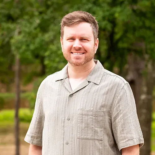 Man in tan shirt smiles outdoors in front of trees.