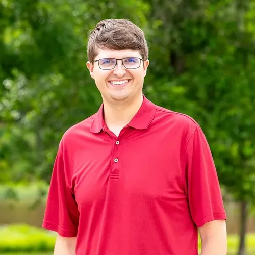 Man with glasses smiling in red polo shirt, in front of green trees.
