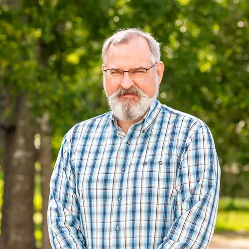 Man with a graying beard wearing glasses and a blue plaid shirt, outdoors in front of trees.