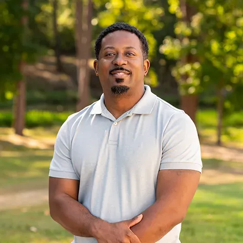 Man with dark skin smiles, wearing a gray polo shirt, standing outdoors with trees in the background.