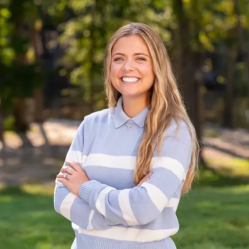 Blonde woman smiles, arms crossed. Wearing light blue sweater with white stripes, outdoor setting.