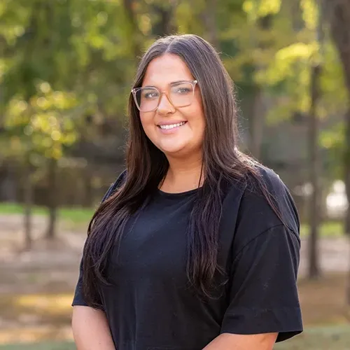 Woman with long brown hair, glasses, and black shirt smiling in an outdoor setting.