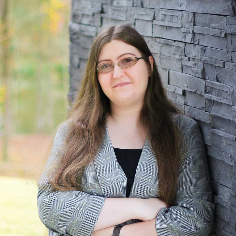 A woman wearing glasses is leaning against a brick wall with her arms crossed.