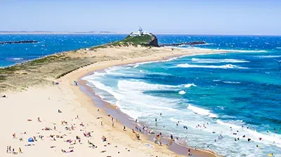 A sandy beach with people swimming and relaxing, leading to a scenic headland with a white lighthouse under a clear sky.