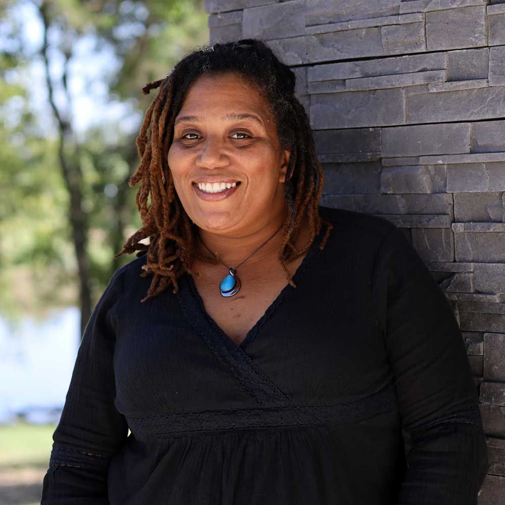 A woman with dreadlocks is leaning against a stone wall and smiling.