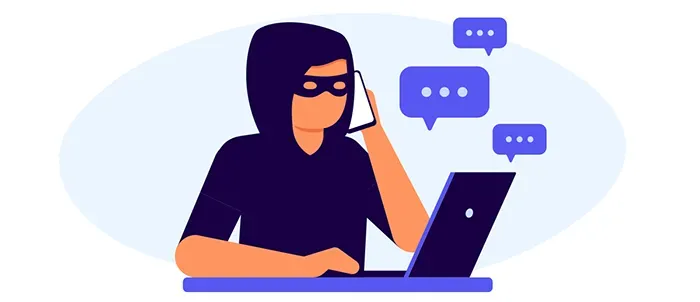 A man in a hood is talking on a cell phone while using a laptop computer.