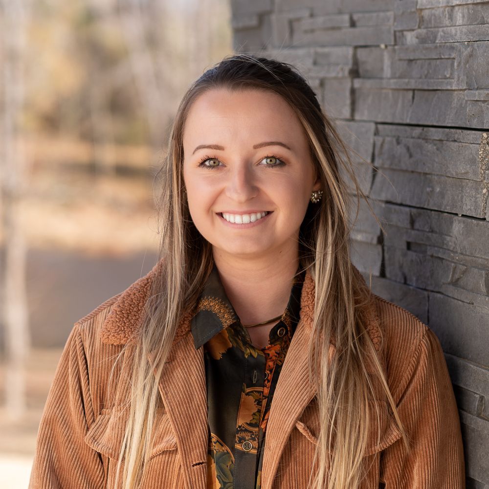 A woman in a brown jacket is smiling while leaning against a stone wall.