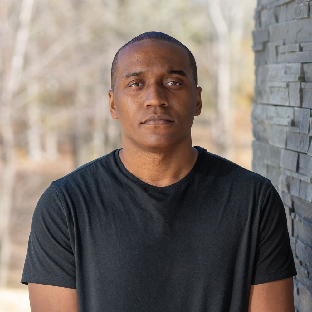A man in a black shirt is standing in front of a stone wall.