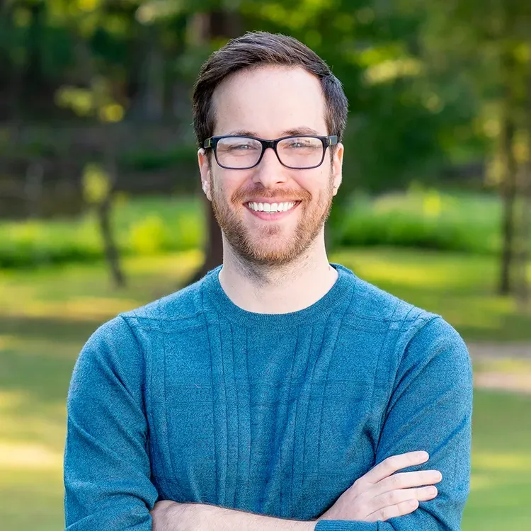 A man wearing glasses and a blue sweater is smiling with his arms crossed.