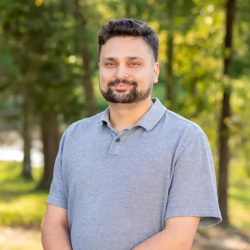 A man with a beard is wearing a grey polo shirt and standing in front of trees.