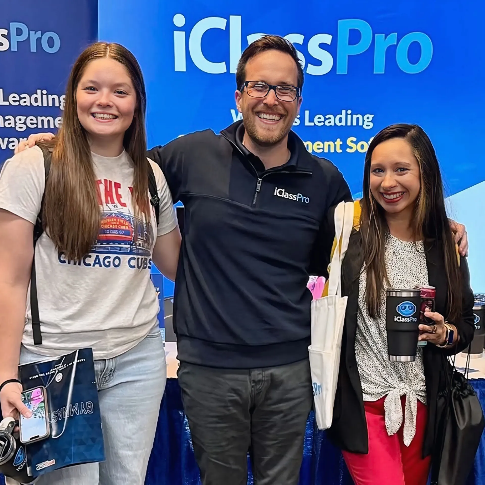 Three people smiling in front of a blue iClassPro promotional backdrop at a conference.