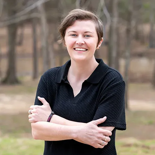 A woman in a black polo shirt is smiling with her arms crossed.
