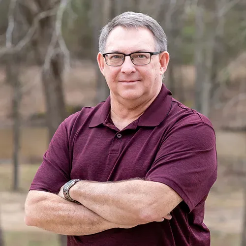 Man with glasses and crossed arms, wearing a maroon polo, outdoors.