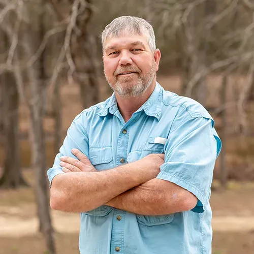 A man in a blue shirt is standing with his arms crossed in front of trees.