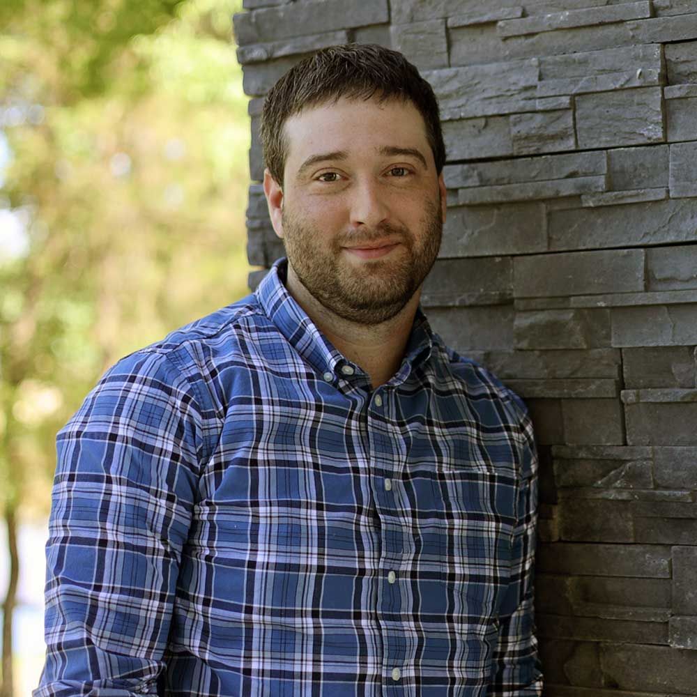 Man in blue plaid shirt leans against a gray stone wall, smiling. Outdoors.