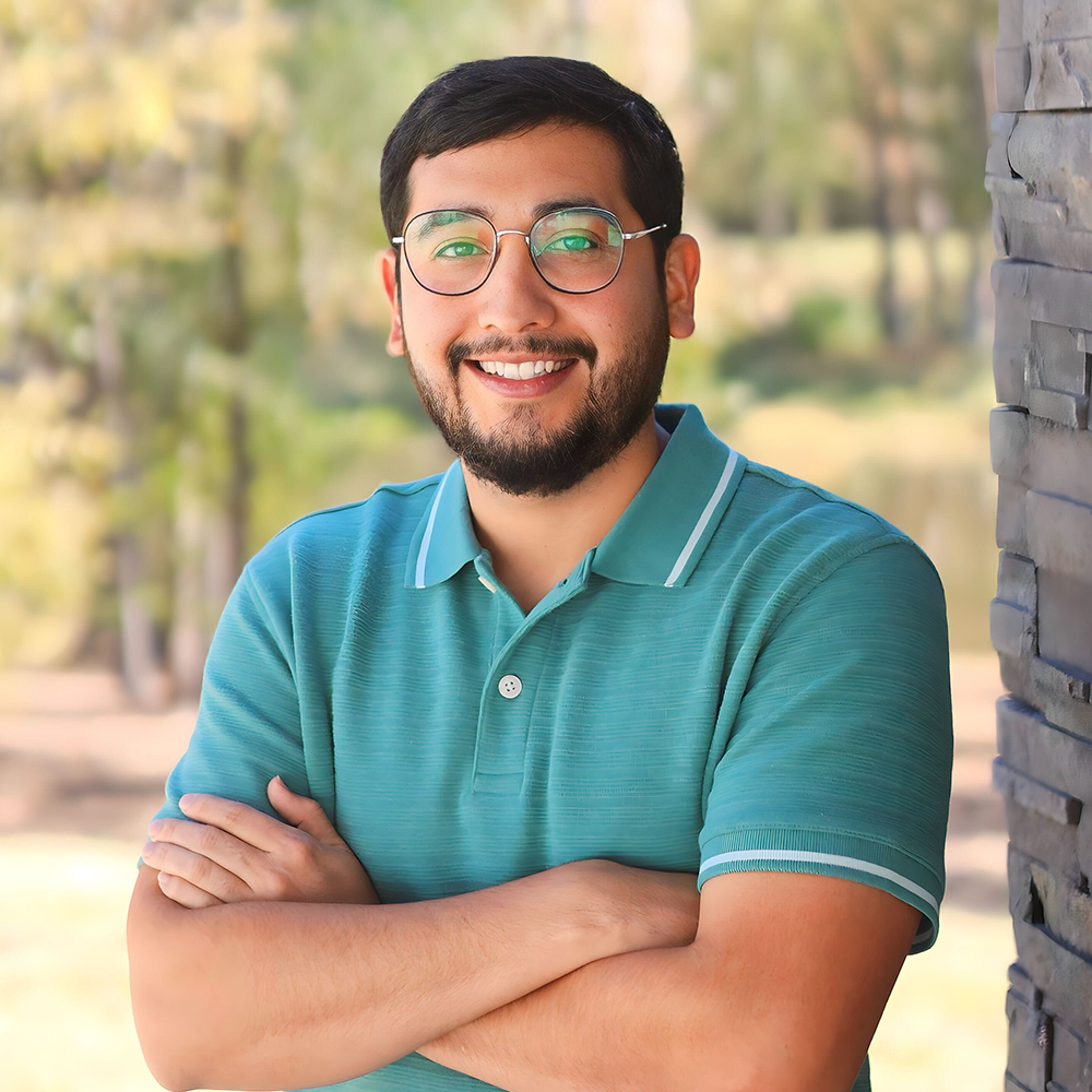 A man wearing glasses and a green polo shirt is smiling with his arms crossed.