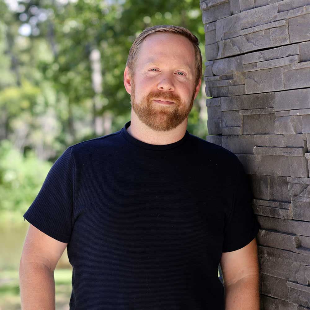 A man with a beard is leaning against a stone wall