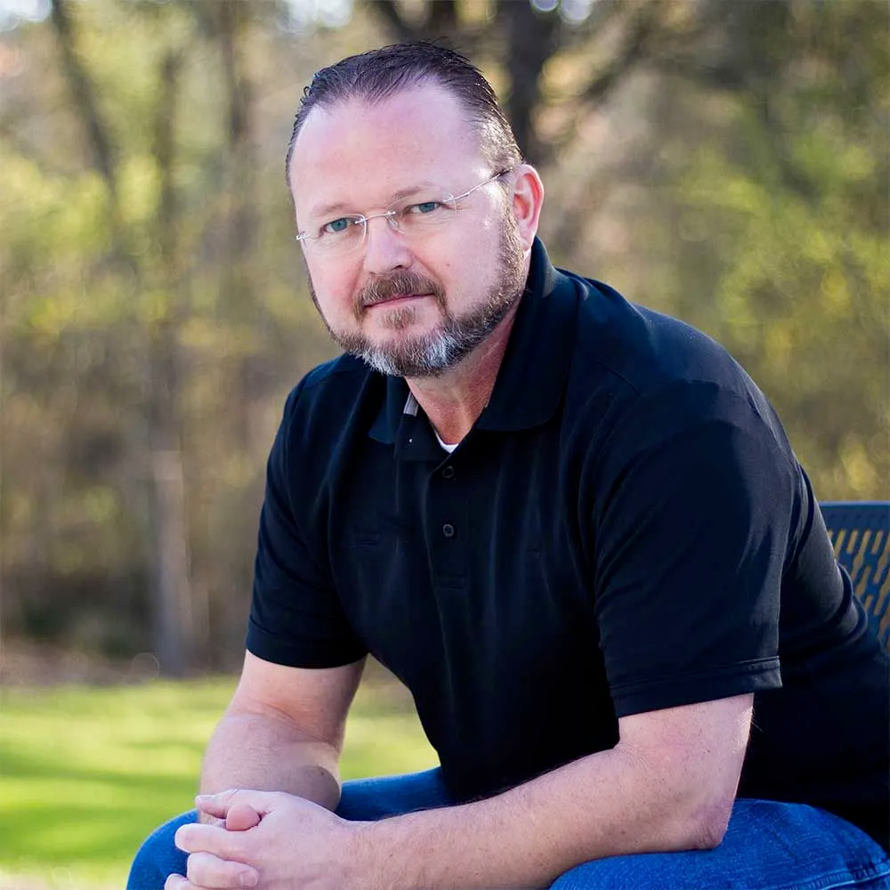 A man with glasses and a beard is sitting on a park bench.