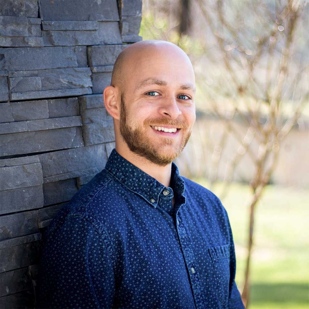 A bald man with a beard is leaning against a stone wall and smiling.