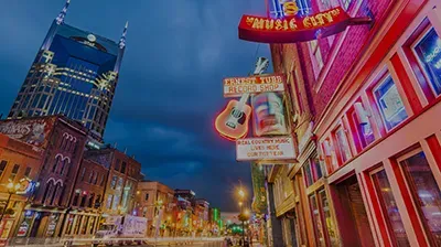 A neon-lit Nashville street at night, featuring historic bars with vibrant signs and the tall AT&T Building in the back.