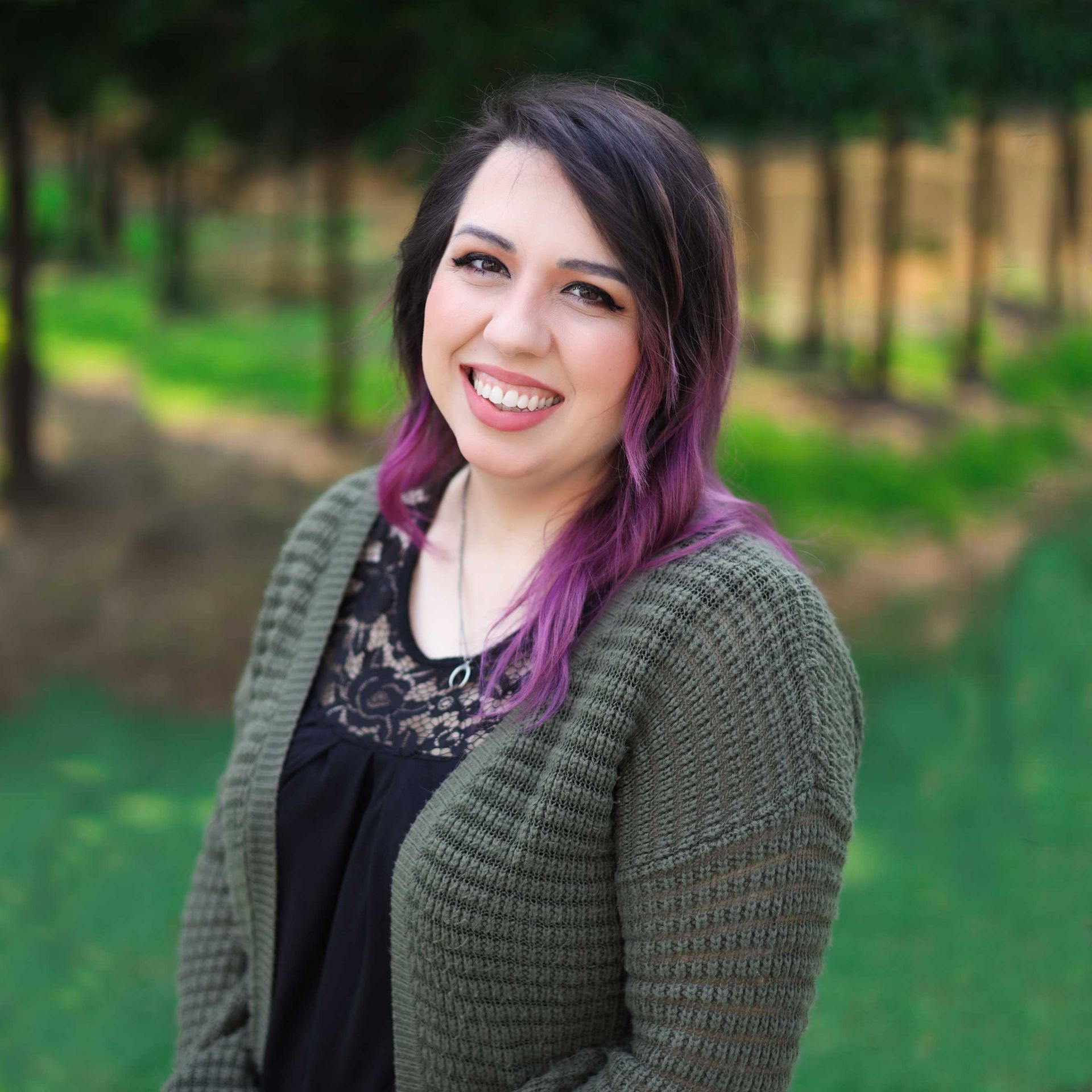 A woman with purple hair and a green sweater is smiling for the camera.