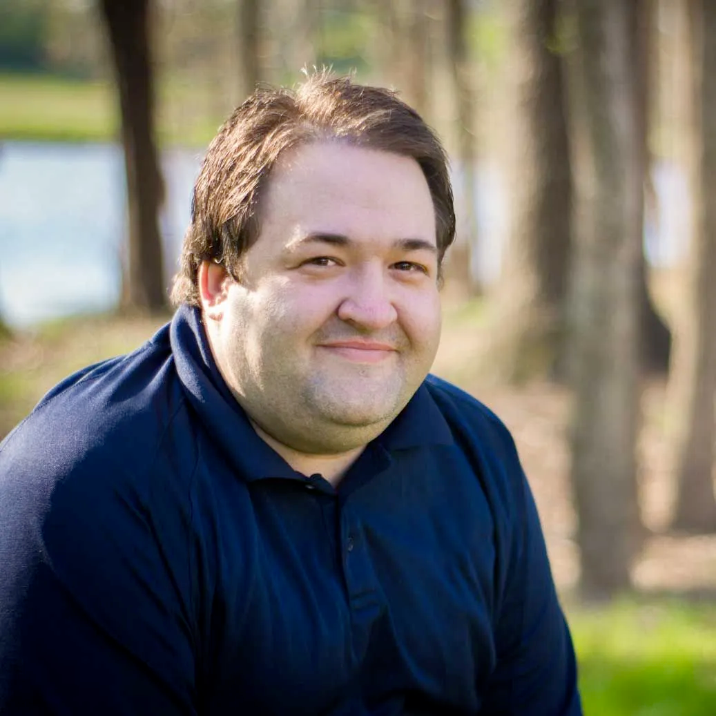 A man in a blue shirt is sitting in the grass and smiling for the camera.