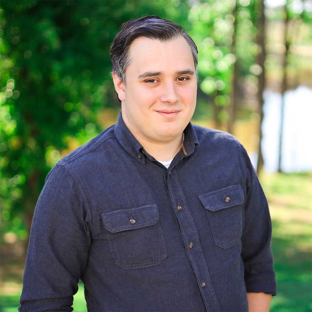 A man in a blue shirt is standing in front of trees.