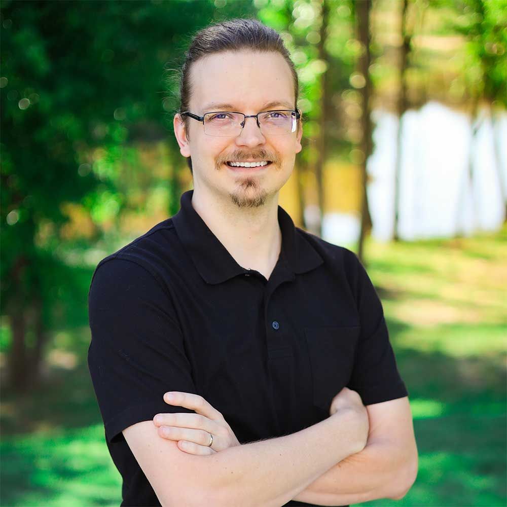 A man wearing glasses and a black shirt is standing with his arms crossed and smiling.