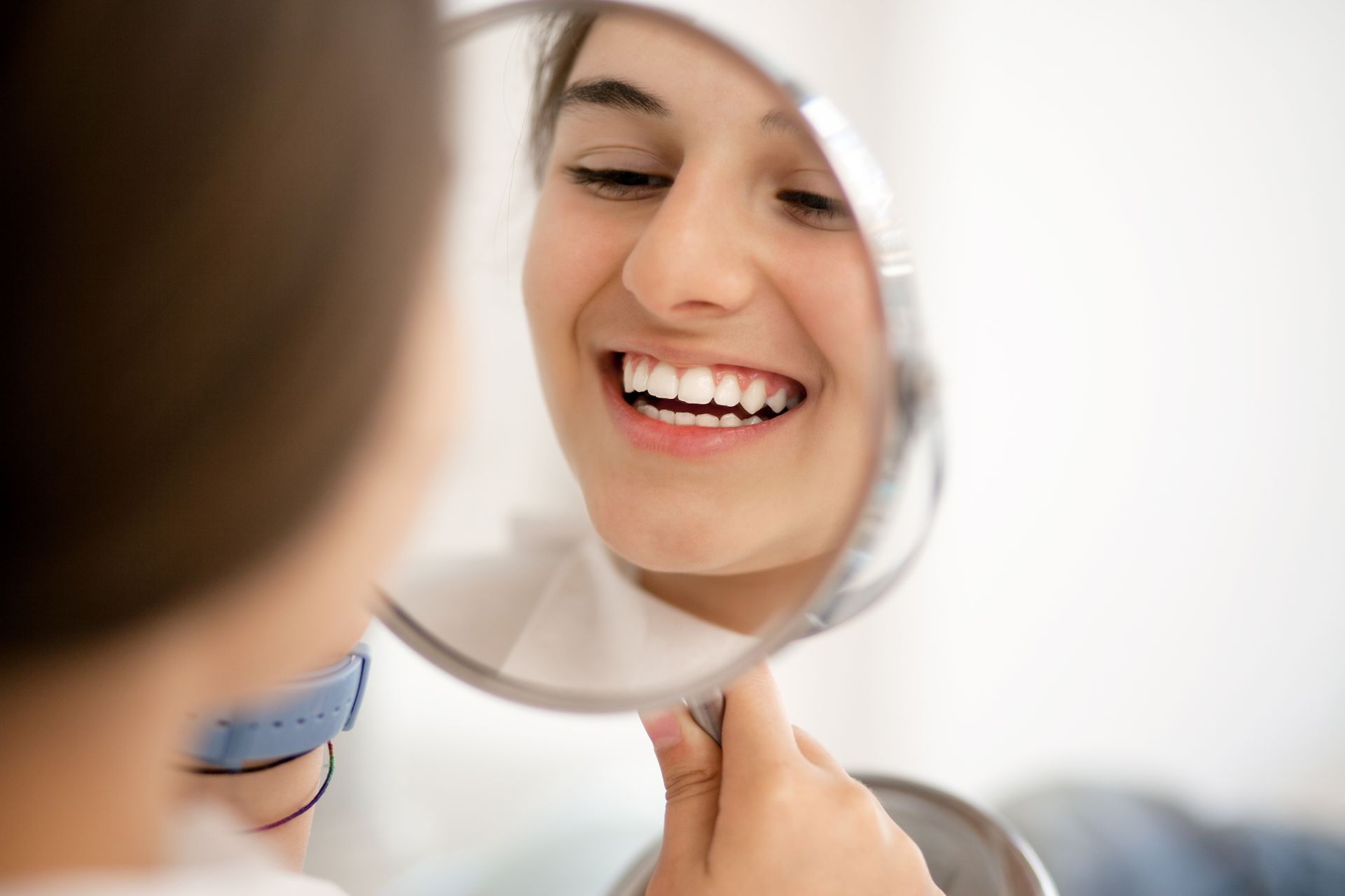 Woman smiling, looking at her teeth in a hand mirror. Indoors, natural light.