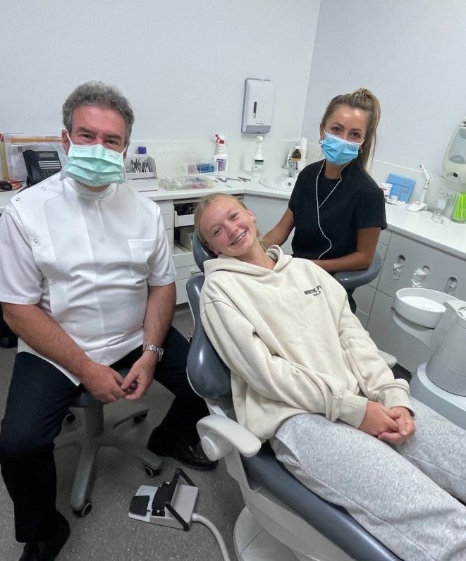 Dentist and assistant with patient in dental chair. All wearing masks. Smiling patient, white room.