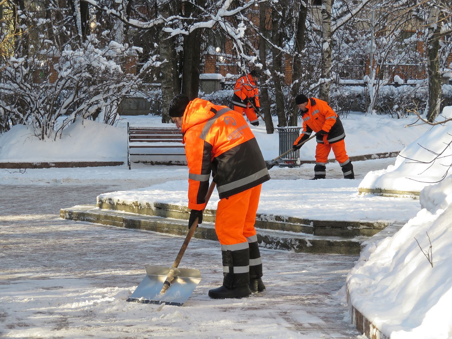 Workers in bright orange clearing snow from a path in a snowy park