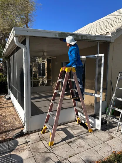 A man is standing on a ladder painting a screened in porch.