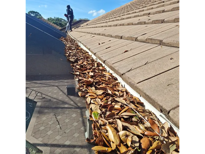 A man is standing on top of a roof with a gutter filled with leaves.