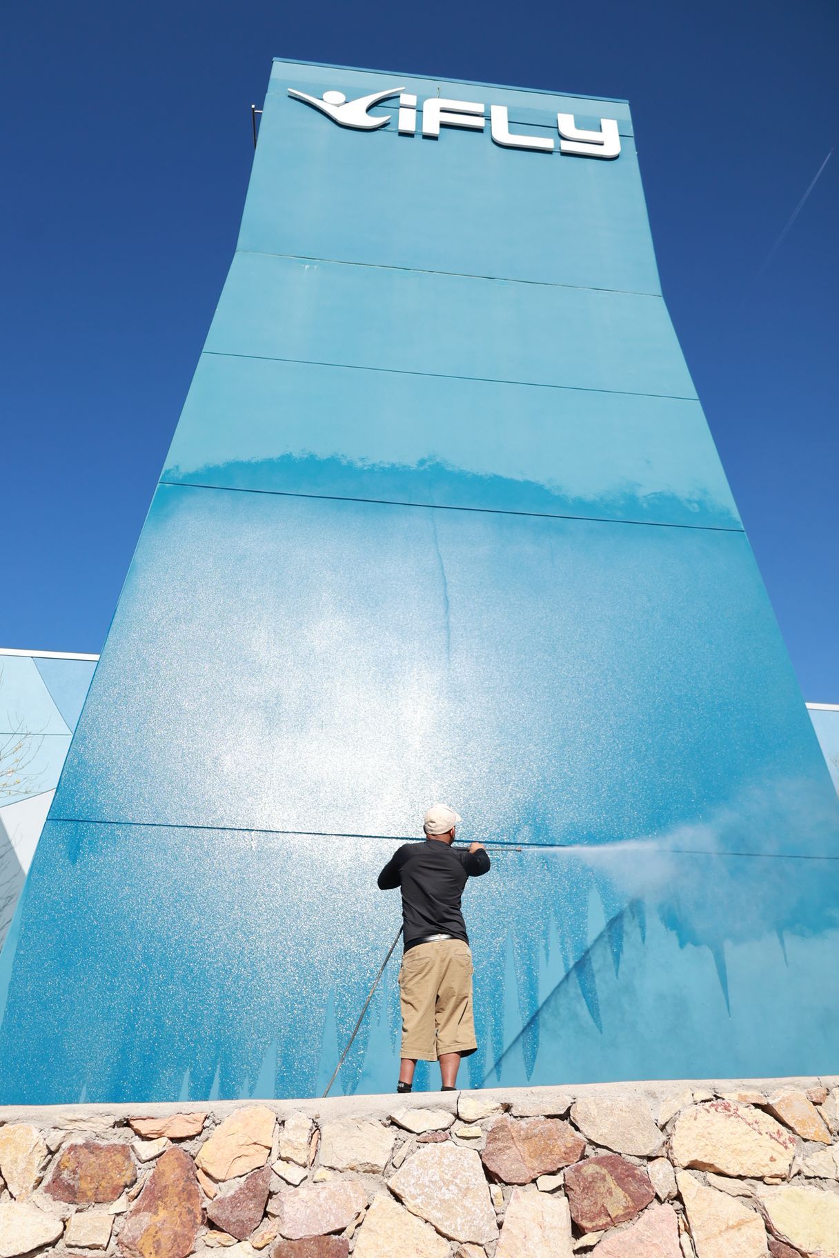 Person standing against a tall blue IFly climbing wall under a clear sky.