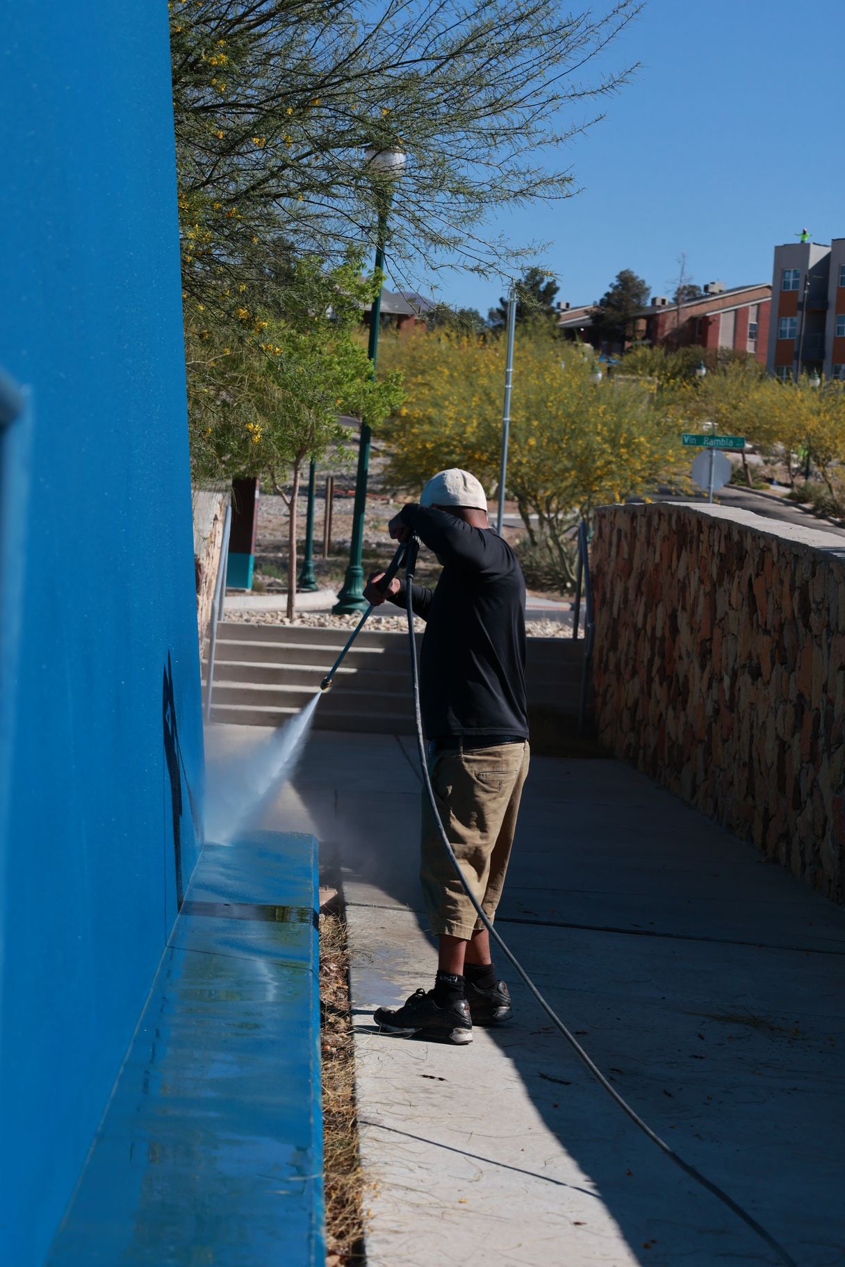 Person in dark clothing stands near a blue wall on a sunny sidewalk, looking out over a stone-lined path.