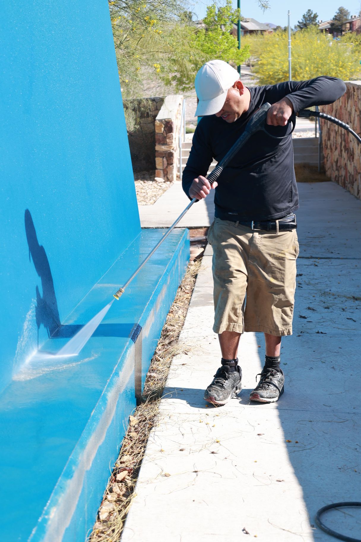 Person pressure washing a bright blue wall on a sunny outdoor walkway.