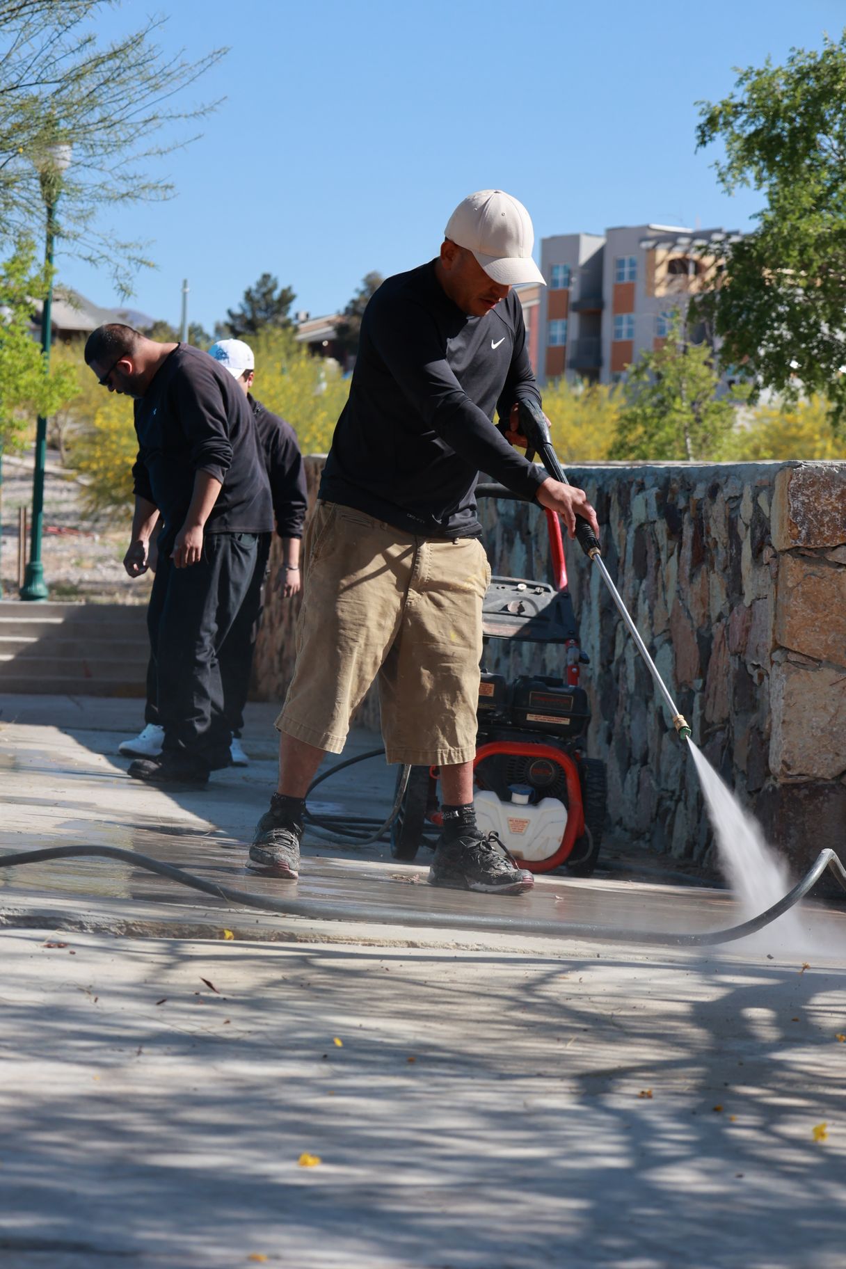 Two workers pressure-washing a sidewalk beside a stone wall on a sunny day