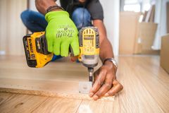 Person using a yellow power drill to attach a small, white hardware piece to a wooden board on a light wooden floor.