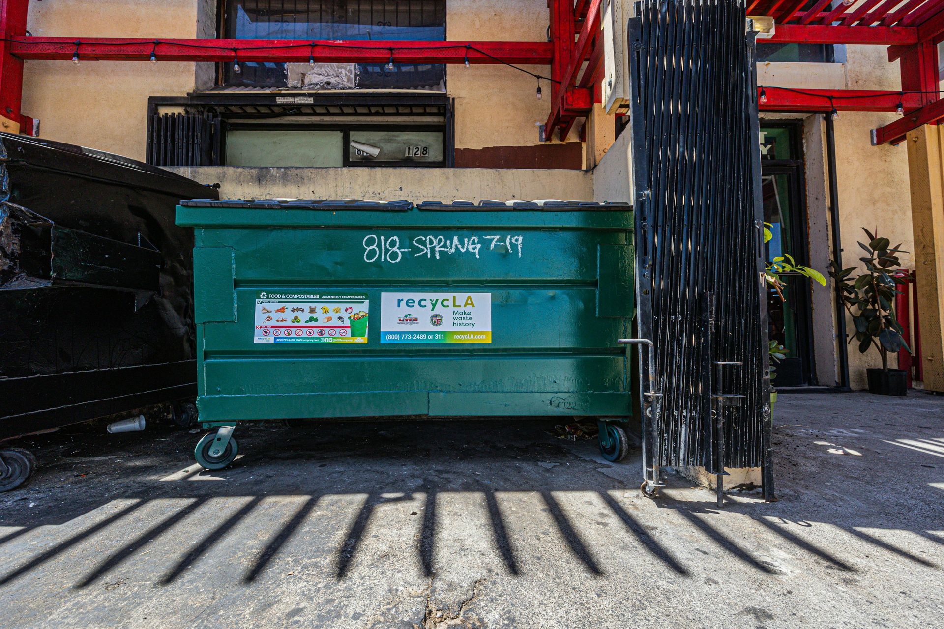 Green dumpster in a courtyard with red beams and a black tree trunk, casting striped shadows.