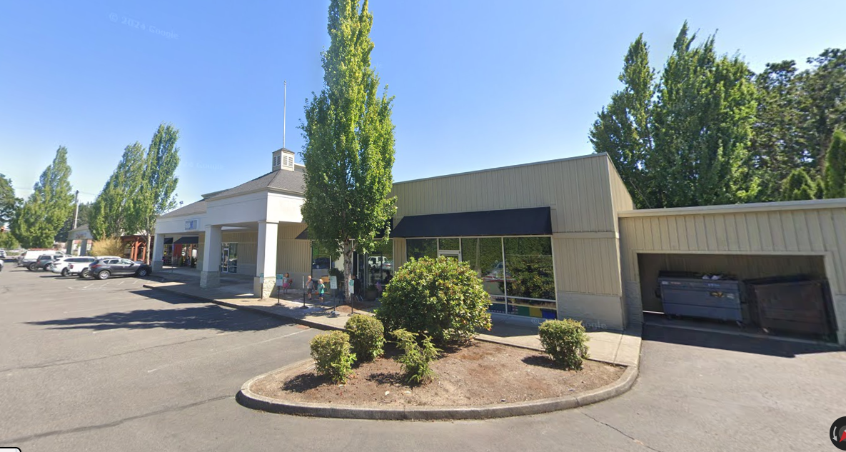 A building with a black awning is surrounded by trees and bushes in a parking lot.