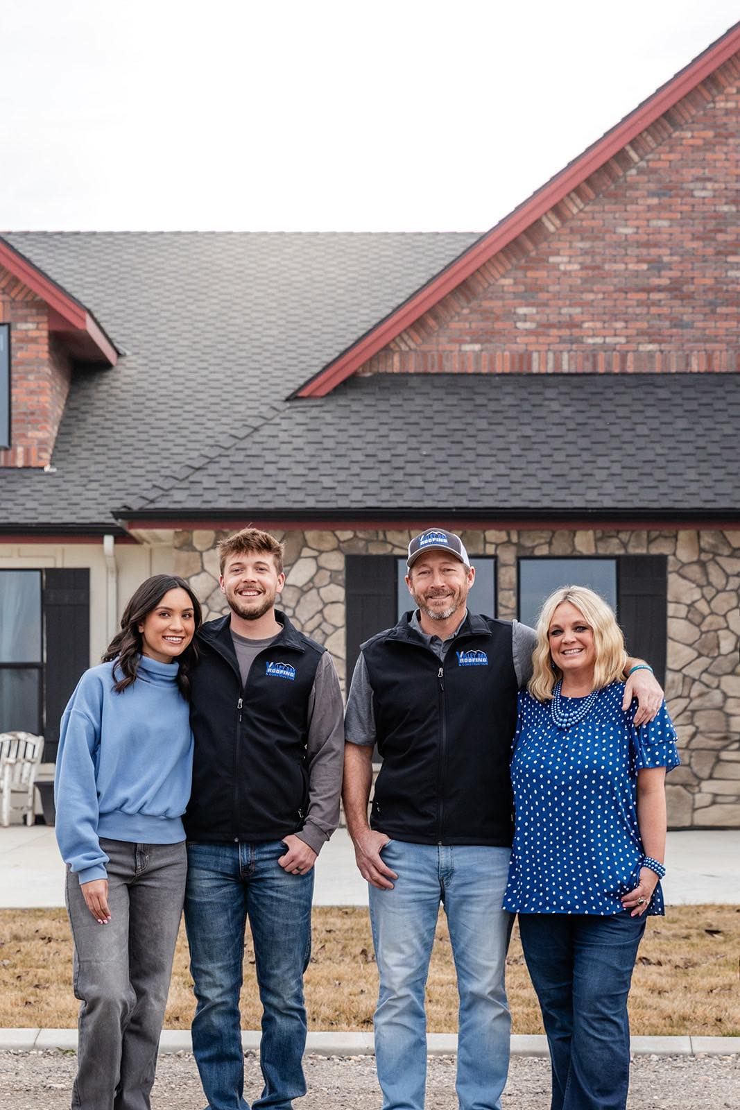 Four people stand smiling in front of a brick house with a shingled roof.
