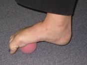 Foot resting on a pink massage ball, for self-treatment on a grey carpet.