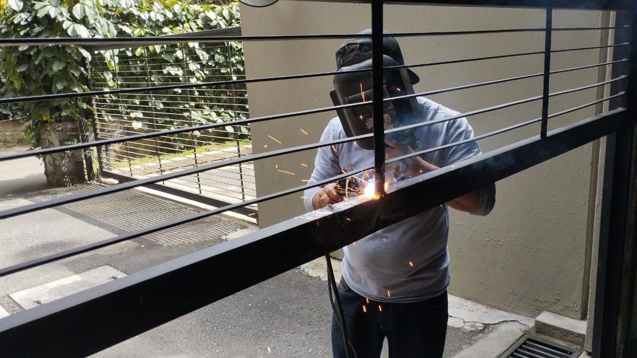 Un trabajador, con un casco de soldadura protector, suelda una puerta metálica horizontal, emitiendo chispas en un entorno al aire libre.