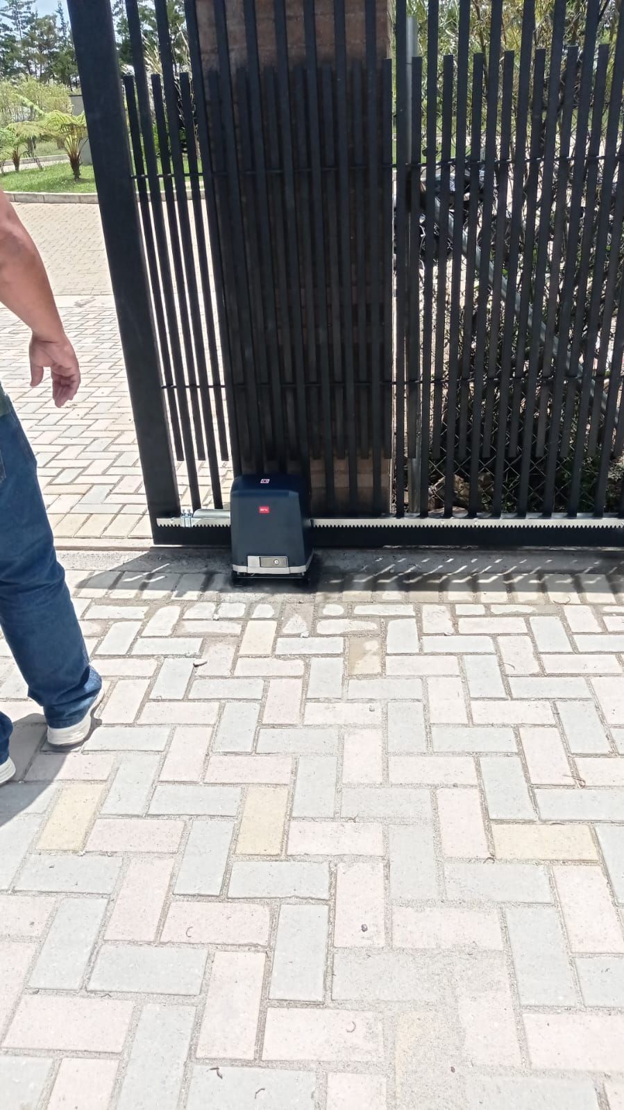 Una persona se encuentra de pie cerca de un abrepuertas motorizado acoplado a una puerta corredera de metal oscuro en un camino de entrada pavimentado.