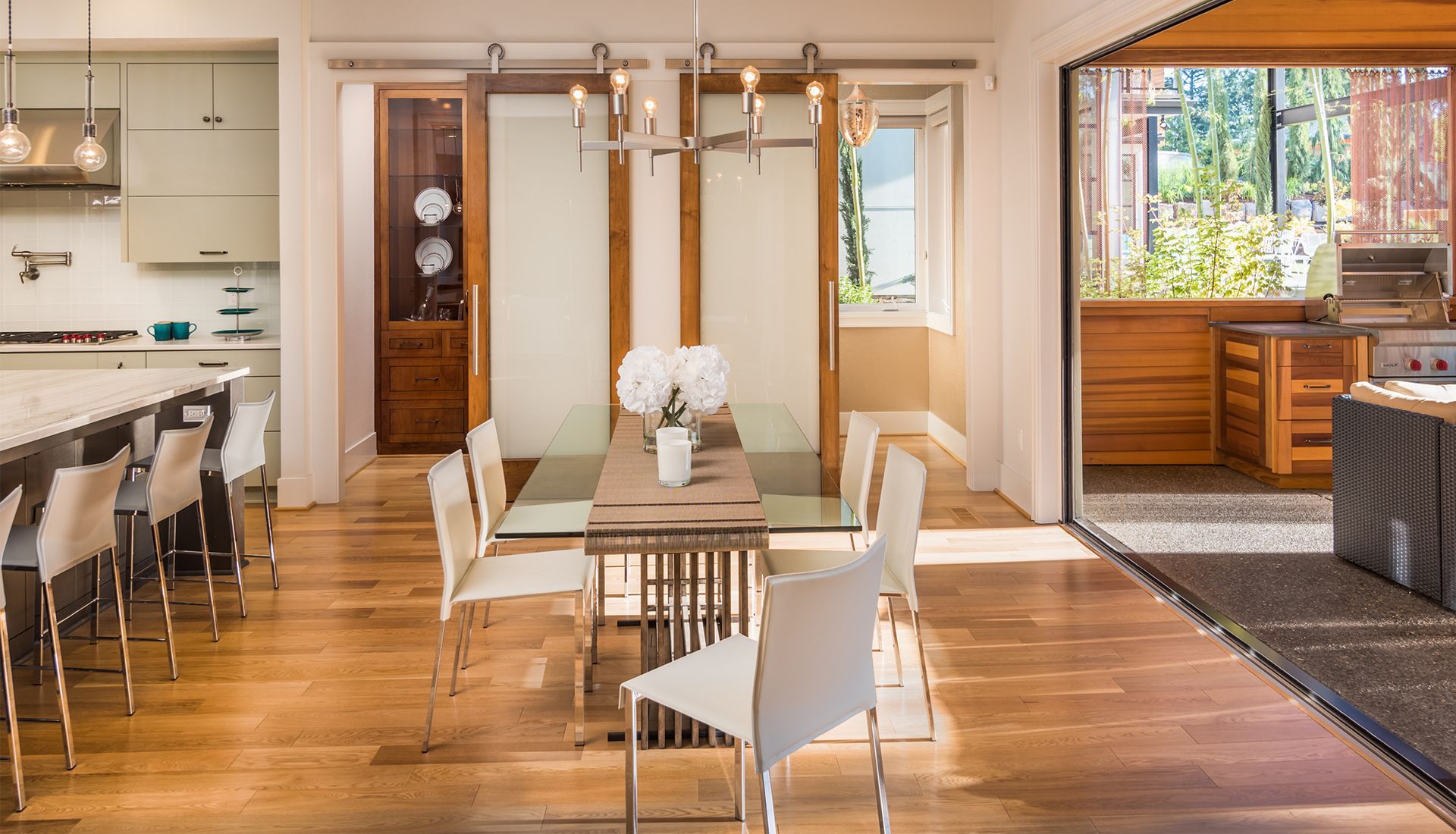 Dining room with glass table, white chairs, and wood floors. Open to a kitchen and outdoor patio.
