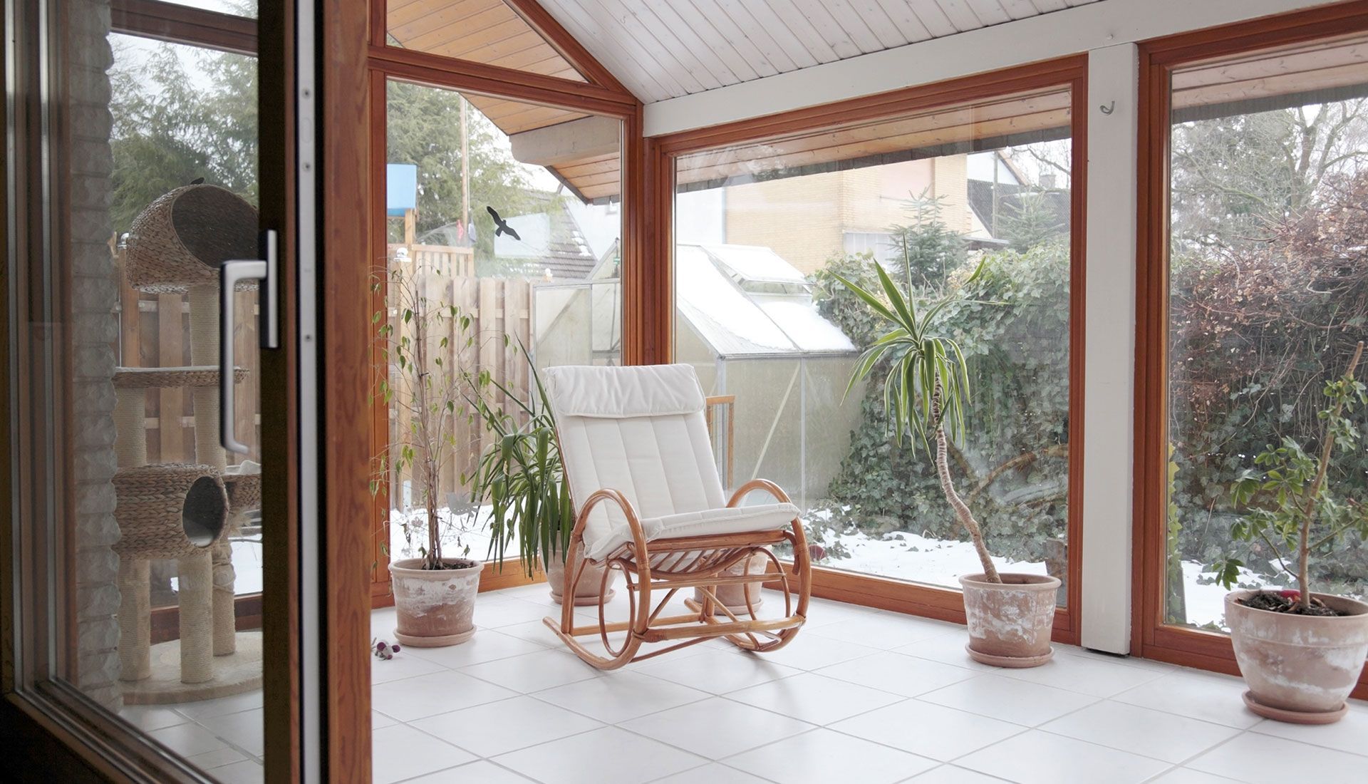 Sunroom with rocking chair, potted plants, and snowy outdoor view through large windows.