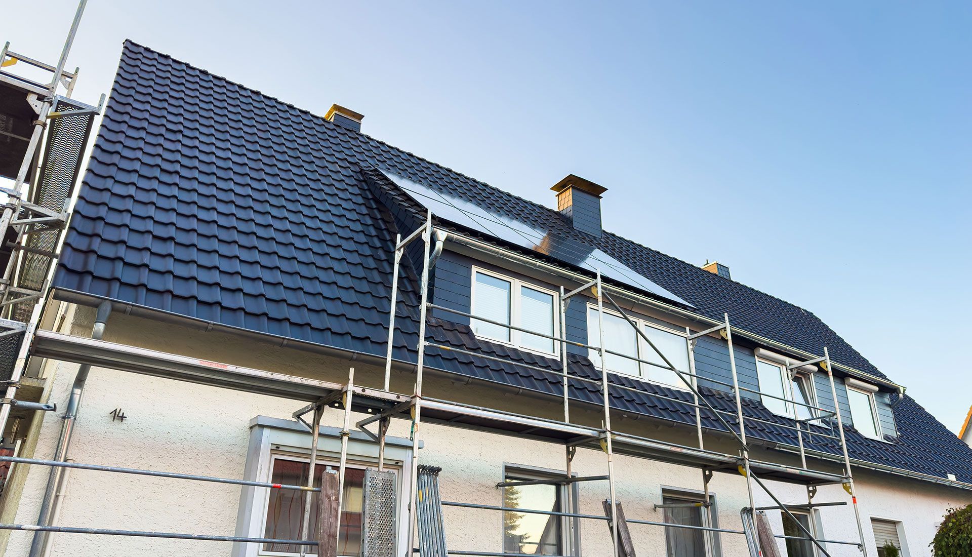 House under construction, new black roof, scaffolding, light-colored facade, windows, clear blue sky.