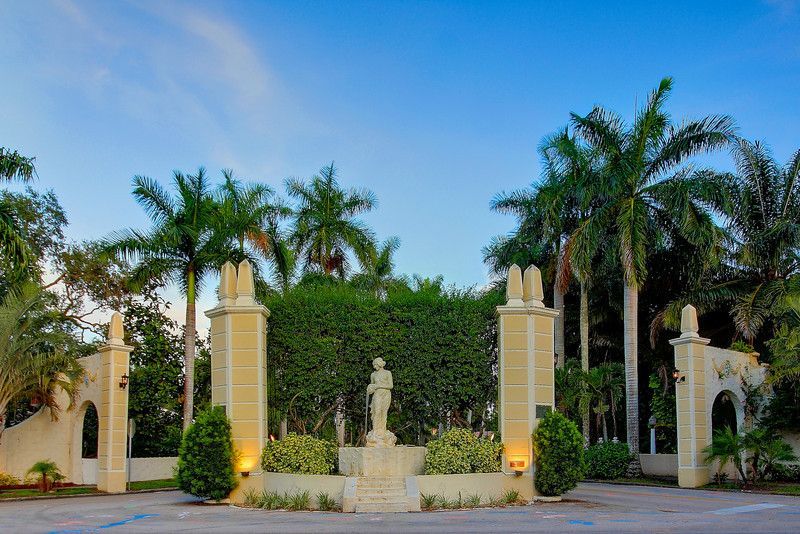 A statue of a man stands in front of a gated entrance surrounded by palm trees.