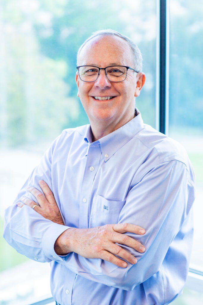 A man in a blue shirt and glasses is standing with his arms crossed in front of a window.