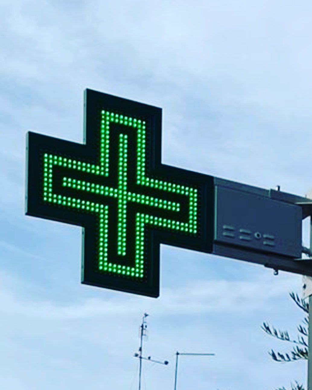 Cartello di farmacia con croce illuminata di verde contro un cielo nuvoloso.